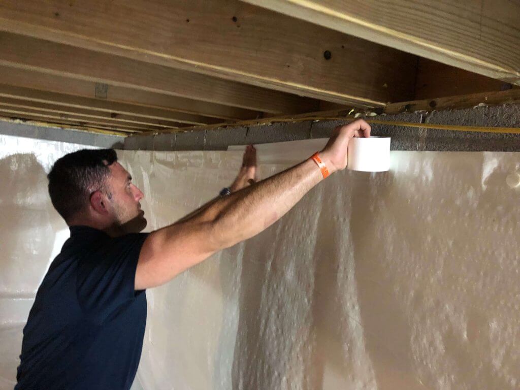 Worker taping a crawl space liner to cinder blocks.