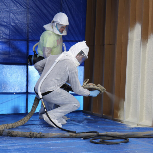 One person demonstrates how to install spray foam insulation while another looks on