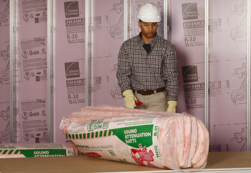 Man in construction hat and glasses wearing gloves and holding a box cutter looking at roll of pink foam