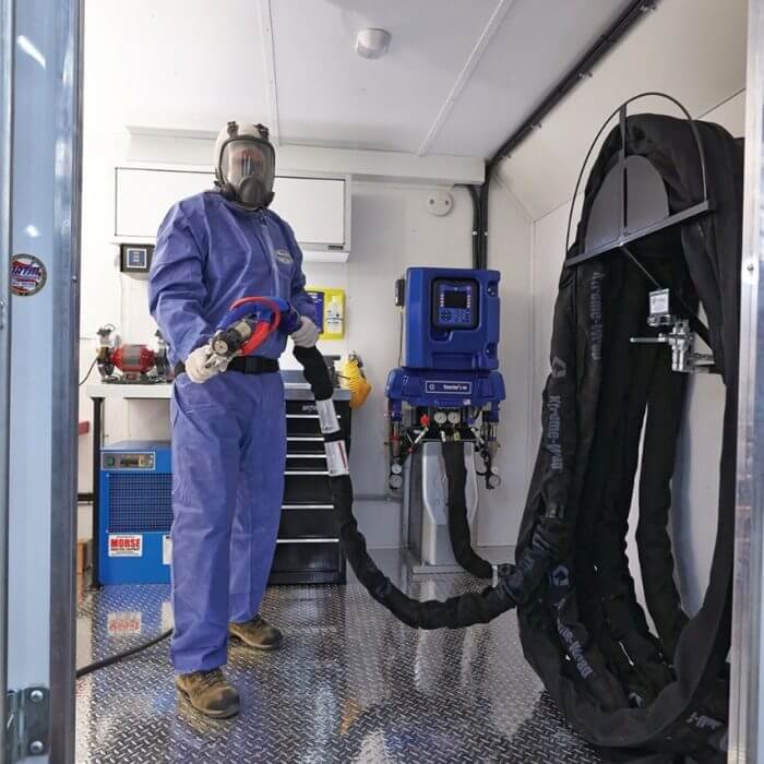 man in coveralls and respirator mask standing inside a foam rig, holding a hose