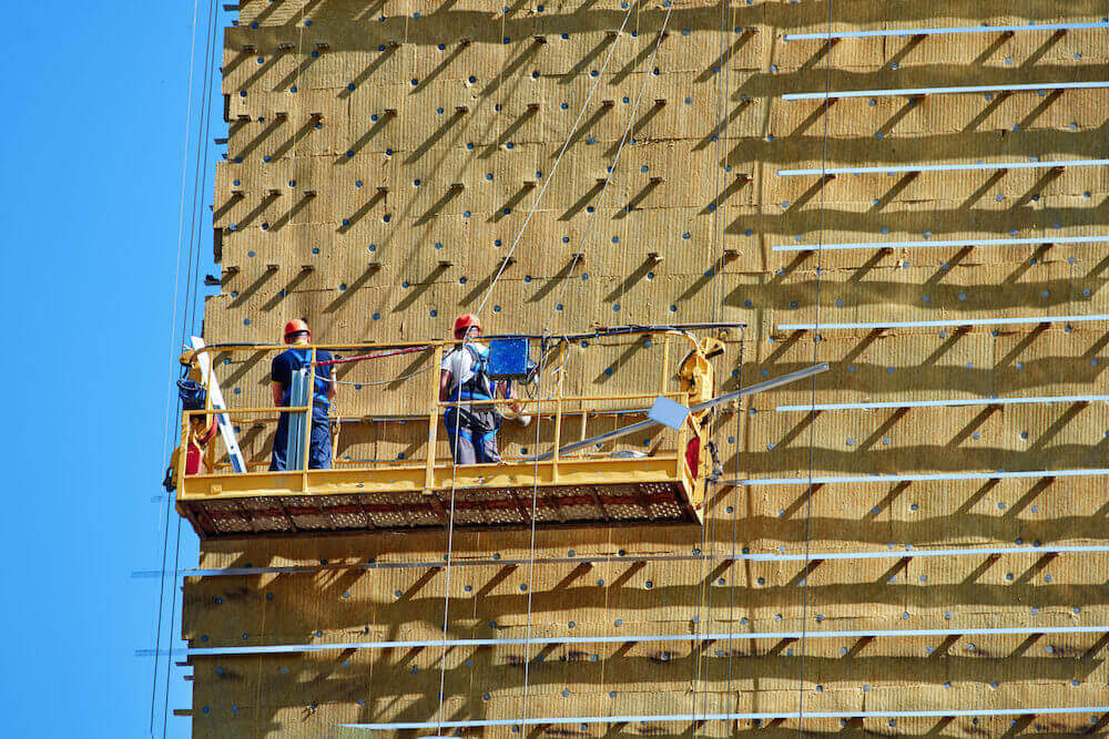 Professional construction Workers on the scaffold elevator insulated wall facade with mineral wool. roofers wearing safety harness insulating wall facade at height on old building in a crane.