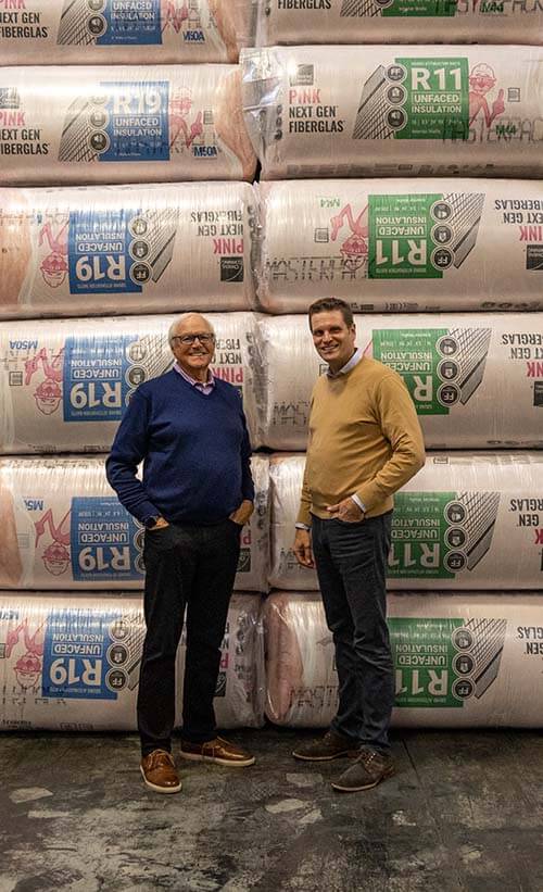 Two smiling, well-dressed men standing in front of large piled stacks of insulation