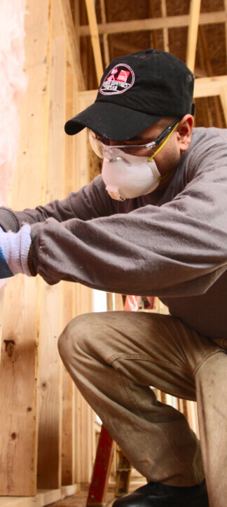A man in a black hat, safety goggles and a white respirator mask working on an unfinished wall.